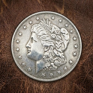 May include: A silver coin featuring a profile of a woman with flowing hair, adorned with a floral crown and the word "LIBERTY". The coin is surrounded by stars and a beaded border, set against a brown leather background.