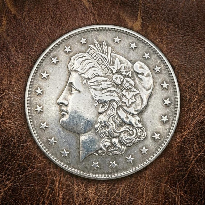May include: A silver coin featuring a profile of a woman with flowing hair, adorned with a floral crown and the word "LIBERTY". The coin is surrounded by stars and a beaded border, set against a brown leather background.