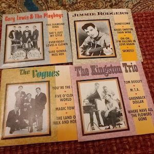 Puede incluir: Se muestran cuatro álbumes de discos de vinilo vintage. Los álbumes presentan fotos en blanco y negro de artistas musicales y títulos de canciones. Los artistas incluyen a Gary Lewis & The Playboys, Jimmie Rodgers, The Vogues y The Kingston Trio.