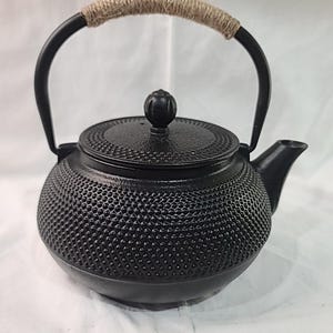 May include: Black cast iron teapot with a textured surface and a woven handle. The teapot has a rounded body, a lid with a small knob, and a spout. The handle is made of black metal with a woven, tan-colored material wrapped around it.