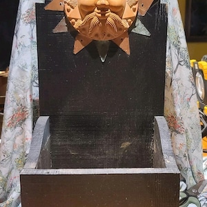 Puede incluir: Una jardinera rústica de madera negra con un diseño de cara de sol. La cara del sol está hecha de terracota y tiene una cara detallada con bigote. La jardinera tiene forma de caja rectangular.