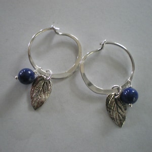 May include: A pair of silver hoop earrings with a small silver leaf charm and a dark blue gemstone hanging from each hoop.