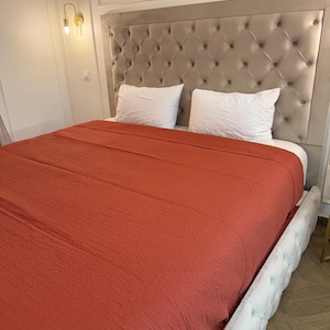 May include: A bed with a tufted headboard and a rust-colored blanket. Two white pillows rest on the bed. The bed frame is white with a button-tufted design. A gold-colored wall sconce is visible on the wall.