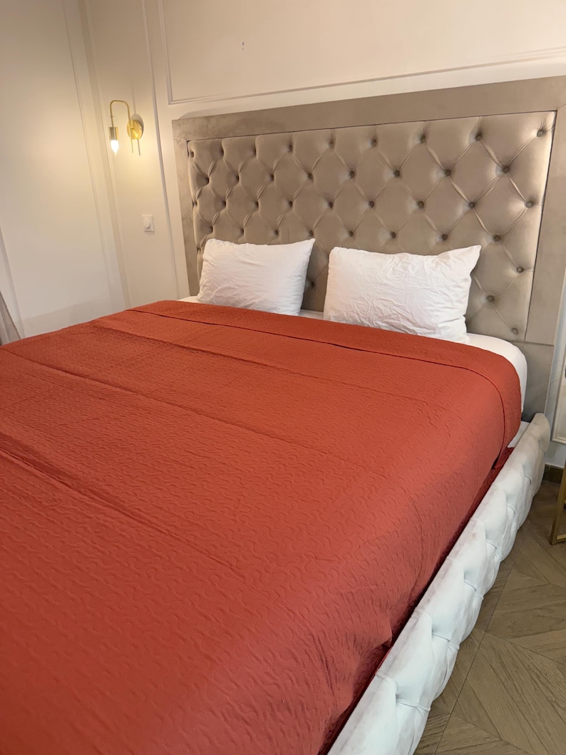 K&ouml;nnte beinhalten: Ein Bett mit einem gesteppten Kopfteil und einer rostfarbenen Decke. Zwei wei&szlig;e Kissen liegen auf dem Bett. Der Bettrahmen ist wei&szlig; mit einem Knopf-getufteten Design. Eine goldfarbene Wandleuchte ist sichtbar.
