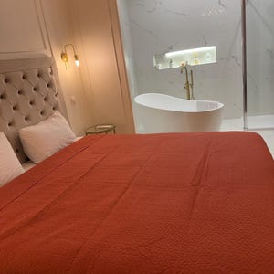 K&ouml;nnte beinhalten: Ein Schlafzimmer mit einem gro&szlig;en Bett, das mit einer rostfarbenen Steppdecke bedeckt ist. Das Kopfteil ist mit einem hellbeigen Stoff gepolstert. Im Hintergrund ist ein Badezimmer mit einer wei&szlig;en Badewanne und Fliesen in Marmoroptik zu sehen.