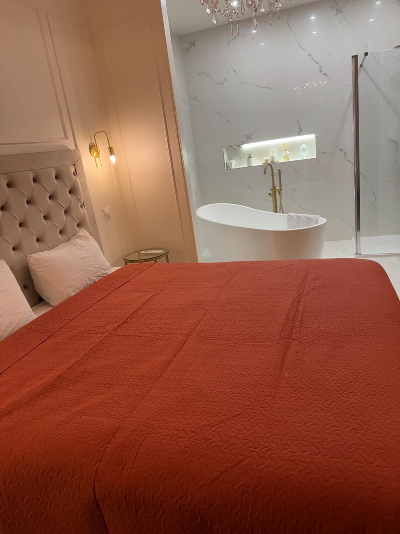 K&ouml;nnte beinhalten: Ein Schlafzimmer mit einem gro&szlig;en Bett, das mit einer rostfarbenen Steppdecke bedeckt ist. Das Kopfteil ist mit einem hellbeigen Stoff gepolstert. Im Hintergrund ist ein Badezimmer mit einer wei&szlig;en Badewanne und Fliesen in Marmoroptik zu sehen.