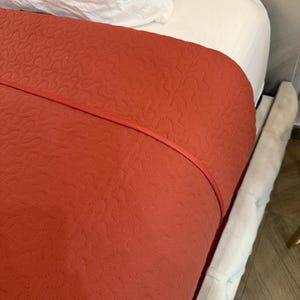 K&ouml;nnte beinhalten: Eine terrakottafarbene, gesteppte Tagesdecke mit roter Paspel, drapiert &uuml;ber einem wei&szlig;en Laken und Kissen. Die Tagesdecke weist ein sich wiederholendes, gen&auml;htes Muster auf. Der Bettrahmen ist mit einem hellgrauen Stoff bezogen.