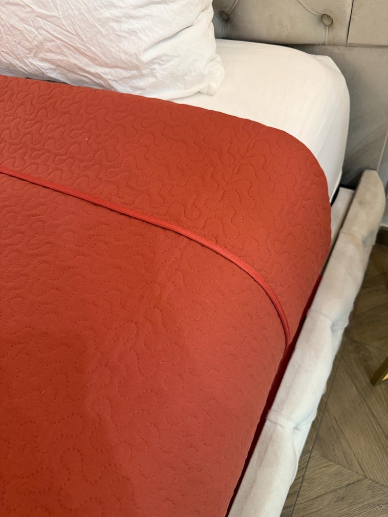 K&ouml;nnte beinhalten: Eine terrakottafarbene, gesteppte Tagesdecke mit roter Paspel, drapiert &uuml;ber einem wei&szlig;en Laken und Kissen. Die Tagesdecke weist ein sich wiederholendes, gen&auml;htes Muster auf. Der Bettrahmen ist mit einem hellgrauen Stoff bezogen.