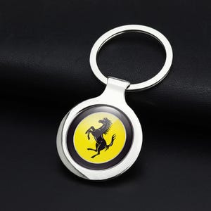 Può includere: Un portachiavi circolare in metallo argentato con un emblema centrale su sfondo giallo raffigurante la sagoma di un cavallo nero rampante. Il design è racchiuso in una cornice d'argento lucido e fissato a un anello metallico.