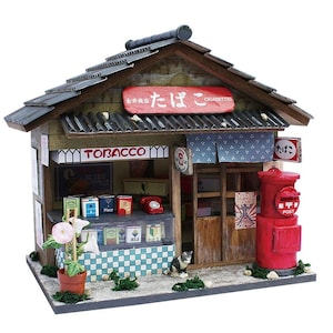Puede incluir: Modelo en miniatura de una tabaquería japonesa con un buzón rojo y un pequeño gato. La tienda tiene una fachada de madera, un tejado de tejas y letreros que dicen "TOBACCO" y "CIGARETTES". La tienda también tiene una vitrina con varios artículos.