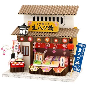 May include: A miniature Japanese shop model made from corrugated cardboard. The shop has a sign with Japanese text, a lantern, and a display of packaged goods. It is decorated with paper flowers and a small sign, evoking a traditional aesthetic.