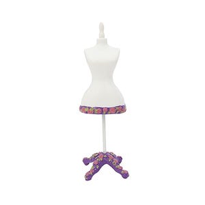 Puede incluir: Un pequeño maniquí blanco con un borde floral en la base. Está sostenido por un poste blanco y una base morada en forma de flor. La base y el borde presentan flores rosas y amarillas.