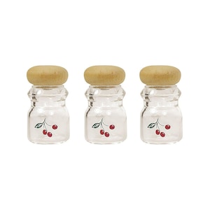 May include: Three clear glass jars with wooden lids. Each jar has a cherry design on the front. The cherries are red with green leaves. The jars are small and round, perfect for storing spices or small items.