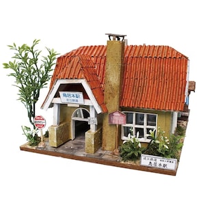 May include: A miniature model of a Japanese train station with a red tile roof, a chimney, and a sign that reads "Torimoto Station". The model includes a small tree and other miniature plants. The station has a white trim and a sign that reads "Omi Railway".