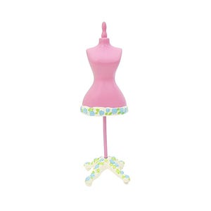 Puede incluir: Un maniquí rosa en miniatura con un borde floral decorativo en la parte inferior. El maniquí está sostenido por un fino poste rosa y una base blanca con detalles florales azules. El maniquí es un artículo decorativo.