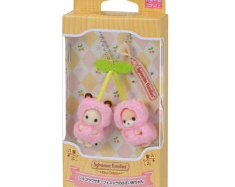 May include: A Sylvanian Families cherry-themed keychain featuring two small animal figures wearing pink fuzzy hooded outfits. The figures are attached to a silver ball chain and green fabric leaves, all housed in a rectangular cardboard box with a clear window and Japanese text.