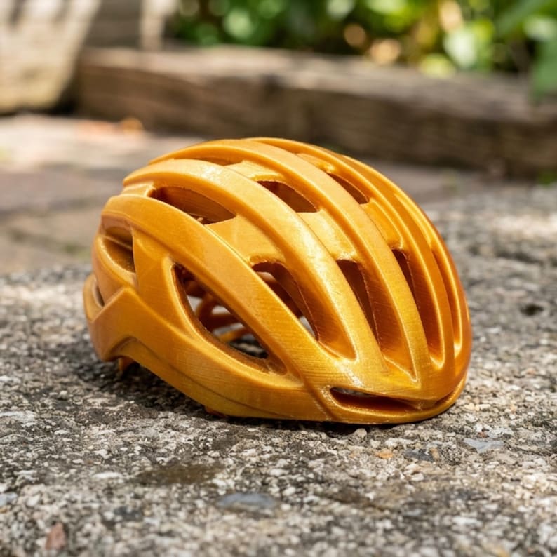 May include: A golden-yellow bicycle helmet with multiple ventilation slots. The helmet is made of a lightweight material and is designed for safety and comfort. The helmet is sitting on a textured, gray surface.