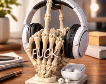 May include: A bone-colored skeleton hand-shaped headphone stand holding white and black headphones. The word "FUCK" is displayed on the hand. A white charging case with earbuds and two additional earbuds are on the base.