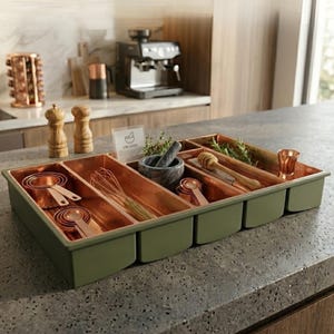 Peut inclure: Un organisateur de cuisine vert avec des compartiments doublés de cuivre. Il contient des tasses à mesurer, un fouet, un mortier et un pilon, une cuillère à miel et une petite tasse en cuivre. L'organisateur est posé sur un plan de travail gris.