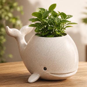 Peut inclure: Un pot de fleurs en forme de baleine en céramique mouchetée, contenant une plante verte vibrante. La baleine a un visage souriant, une queue et une nageoire. Le pot est beige clair et repose sur une surface en bois.