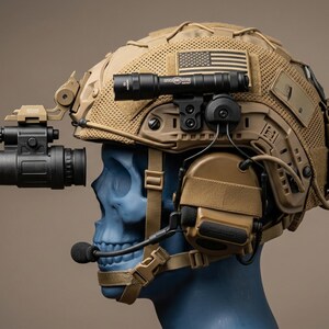 May include: Tan tactical helmet with attached night vision device, flashlight, and communication headset. The helmet features an American flag patch and various mounting accessories. The helmet is displayed on a blue skull.
