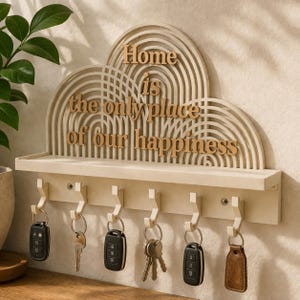 May include: A cream-colored key holder with a shelf and six hooks. The key holder has a decorative arched design with the words "Home is the only place of our happiness" in wooden letters. Keys and key fobs hang from the hooks.