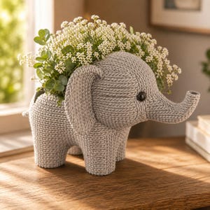Peut inclure: Un pot de fleurs en forme d'éléphant tricoté gris, rempli de fleurs blanches et de feuillage vert. L'éléphant a de grandes oreilles, une trompe et un petit œil noir. Le pot est posé sur une surface en bois.