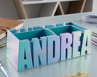 Puede incluir: Un organizador de escritorio personalizado en color turquesa iridiscente, que deletrea "ANDREA" en letras grandes. El organizador tiene dos compartimentos y una pequeña placa dorada con la inscripción "ANDREA - Custom Series". Se ven bolígrafos y cuadernos en el fondo.