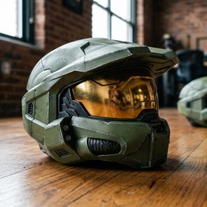 May include: A detailed, olive green helmet with a gold visor, reminiscent of the Halo video game series. The helmet is resting on a wooden surface, showcasing its intricate design and textured finish. A second helmet is visible in the background.