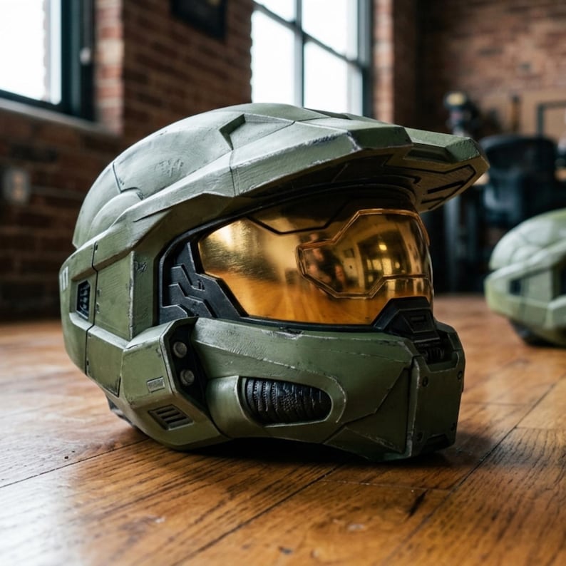 May include: A detailed, olive green helmet with a gold visor, reminiscent of the Halo video game series. The helmet is resting on a wooden surface, showcasing its intricate design and textured finish. A second helmet is visible in the background.