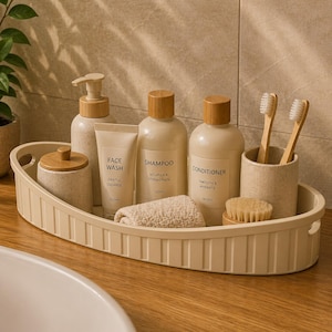 Op de afbeelding: Een beige ovale tray met badkamerbenodigdheden. Het bevat flessen shampoo en conditioner, een tube gezichtsreiniger, een kleine handdoek, een tandenborstelhouder met twee tandenborstels en een kleine borstel. De flessen hebben houten doppen.