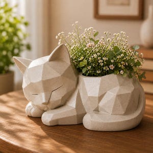Puede incluir: Una maceta con forma de gato beige claro con un diseño geométrico, que contiene un pequeño arreglo de flores blancas y rosas. El gato está acostado con los ojos cerrados, creando una estética pacífica. La maceta está hecha de un material texturizado.
