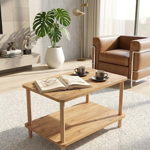 May include: A rectangular light wood coffee table with rounded corners and a lower shelf. The table surface holds an open book with metal-rimmed glasses resting on the pages, alongside two dark gray ceramic mugs on matching saucers.