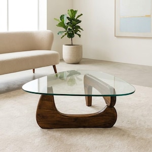 Peut inclure: Une table basse moderne avec un plateau en verre triangulaire et une base en bois foncé. La table a un dessus en verre transparent et une base en bois incurvée. La table est posée sur un tapis de couleur claire dans un salon.