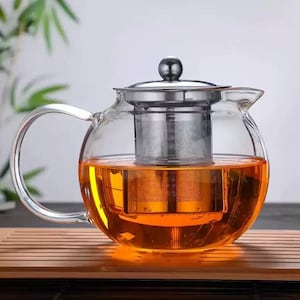 May include: A clear glass teapot with a stainless steel infuser and lid. The teapot is filled with amber-coloured tea. The handle is curved, and the teapot sits on a wooden tray. Bamboo leaves are visible in the background.