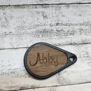 Könnte beinhalten: Ein tropfenförmiges Holzschild mit dem Namen "Abby" in kursiver Schrift graviert. Das Schild hat einen dunklen Rand und ein kleines Loch oben. Der Hintergrund ist eine helle, verwitterte Holzoberfläche.