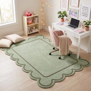 May include: A rectangular area rug in a soft sage green color featuring a scalloped edge design with a darker green border trim. The rug has a plush, solid-colored surface and is positioned on a light-toned wooden floor in a room setting.