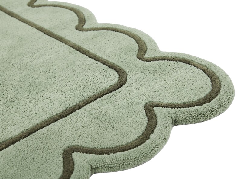 May include: A sage green bath mat with a soft, plush texture and a scalloped edge design. The mat features a darker olive green piped border that follows the curved, wavy outline of the rug, creating a distinct decorative frame around the center.