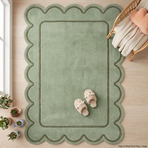 May include: A rectangular sage green plush rug featuring a scalloped edge with a darker green border. The rug has a soft, textured surface and is displayed with a pair of light beige plush slippers resting on its center, showcasing a cozy design.