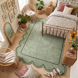 May include: A large, rectangular sage green area rug featuring a scalloped edge border and a darker green inner rectangular frame. The rug is placed on a light wood floor in a bedroom setting, partially tucked under a metal bed frame with floral bedding.