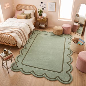 May include: A rectangular area rug in a soft sage green hue featuring a scalloped edge border and a darker green inner rectangular outline. The rug has a plush, solid-colored surface texture and is positioned on a light-toned wooden floor.
