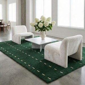 May include: A modern living room setup featuring a deep green rug with white dashes, two white armchairs, and a marble coffee table. A white vase holds a bouquet of white flowers, creating a bright and inviting space.