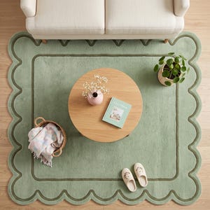 May include: A sage green area rug with a scalloped edge and a dark green border. The rug features a round wooden coffee table, a small potted plant, a book titled Cozy Home, a woven basket with a patterned throw blanket, and two slippers.