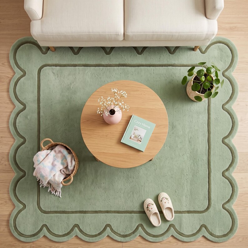 May include: A sage green area rug with a scalloped edge and a dark green border. The rug features a round wooden coffee table, a small potted plant, a book titled Cozy Home, a woven basket with a patterned throw blanket, and two slippers.