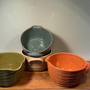 May include: A collection of four ceramic mixing bowls in various colors: olive green, orange, tan, and blue. Each bowl has a unique shape and design, with ribbed detailing and handles. The bowls are arranged on a wooden surface.