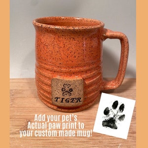 May include: Orange speckled ceramic mug with a brown tile on the side that says "TIGER" with a paw print. The mug has a handle and is sitting on a wooden surface. A black paw print is shown on a white background.