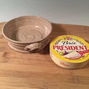 May include: A speckled beige ceramic brie baker with a handle, next to a round package of President Brie cheese. The cheese package is yellow with red and green text and a picture of brie, figs, and grapes.