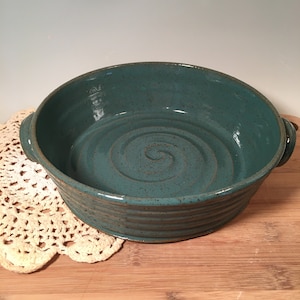 May include: A teal ceramic baking dish with a spiral design in the center. The dish has two handles and a ribbed exterior. It sits on a wooden surface with a cream-colored doily.