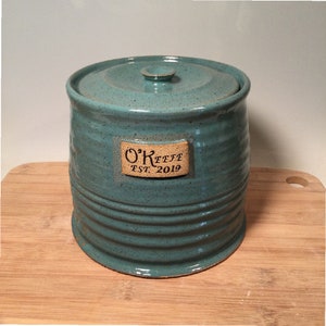 May include: A teal blue ceramic jar with a lid and a brown label that reads "O'Keefe Est. 2019".