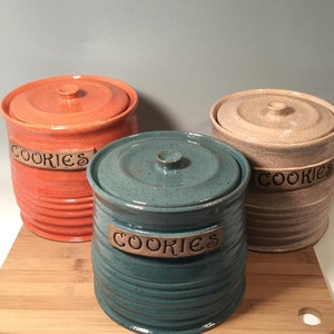 May include: Three ceramic cookie jars with lids. The jars are in different colors: orange, beige, and teal. Each jar has the word "Cookies" written on it.